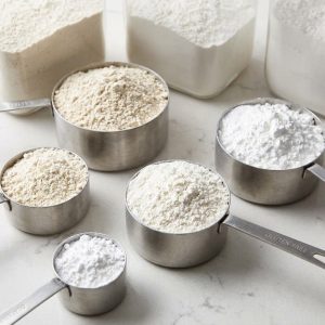 A 1:1 square image showing various gluten-free flours and starches being measured out on a clean kitchen counter, bathed in soft light.