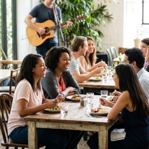A diverse group of friends laugh and dine at rustic tables while a musician performs in the background of a sunlit cafe.